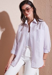 White shirt with purple vertical stripes, button front, two chest pockets, rolled sleeves. Model wearing white pants and sunglasses.