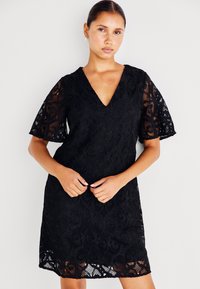 Μαύρο δαντελένιο φόρεμα με V-λαιμόκοψη, φαρδιές κοντές μανίκες και κλος τελείωμα με περίπλοκα λουλουδάτα σχέδια. Το υφασμάτινο υλικό προσθέτει βάθος.