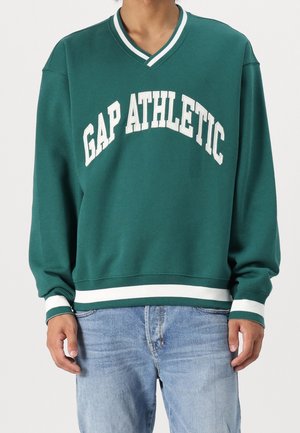 Osoba nosící zelenou mikinu s výstřihem do V, s bílými pruhy a nápisem "GAP ATHLETIC", kombinovanou se světle modrými džínami.
