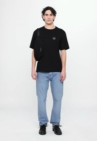 Black cotton t-shirt with a small logo, paired with light blue denim jeans. The model wears a black sling bag and black shoes. White background.