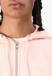 Sweatshirt rosa com fecho éclair, feito de um tecido macio, apresentando um fecho prateado e um logótipo Nike bordado em branco no peito.