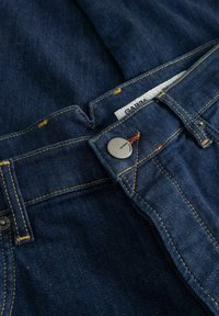 Blugi din denim albastru închis, fața cu nasture metalic și cusături galbene, cu etichetă de marcă parțial vizibilă în interiorul brâului.