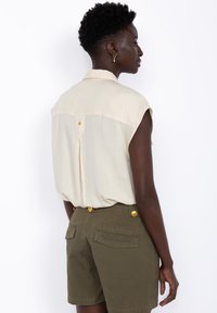 Beige ilmanhiha blouse, jossa on selkäpleat ja tähtikirjonta, yhdistettyinä oliivinvihreisiin shortseihin, joissa on kultaiset napit ja takataskut.