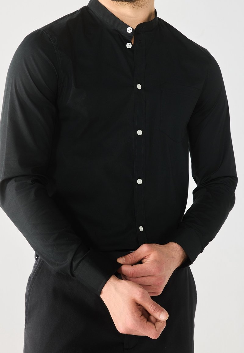 Camicia nera con colletto a band, maniche lunghe, tessuto leggero, taschino anteriore e bottoni argentati lungo l'apertura.