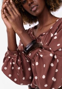 Brązowa bluzka w kropki z bufiastymi rękawami i zapięciem na guziki. Posiada pleciony ciemny bransoletkę i futrzany akcent na nadgarstku.