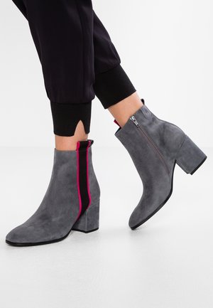 Bottes en daim gris montant à la cheville avec talons blocs, rayure verticale noire et rose, portées avec un pantalon noir retroussé sur fond blanc.