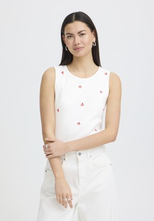 Pikkade tumedate juustega naine, kes kannab valget, varrukateta toppi väikeste punaste lillemustritega ja kõrge vöökohaga valgeid pükse, poseerides käed risti.