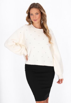 Vrouw in een witte trui met kleine rhinestones en een zwarte kokerrok, poserend met één hand op de heup tegen een witte achtergrond.