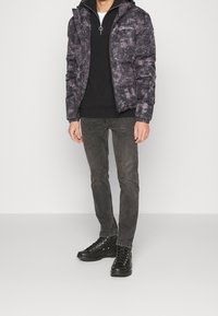 Man som bär en svart och grå tie-dye puffjacka, svart zip-neck tröja, urblekta svarta jeans och svarta high-top sneakers, står mot en vit vägg.