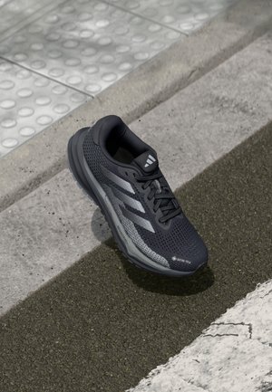 Chaussure de sport noire avec une tige en mesh texturé, accents argentés et une étiquette GORE-TEX. Elle possède une semelle rembourrée et un motif de semelle pour l'adhérence.