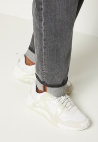 White sneakers with a translucent overlay and textured accents, paired with gray jeans featuring contrasting orange stitching at the cuff.