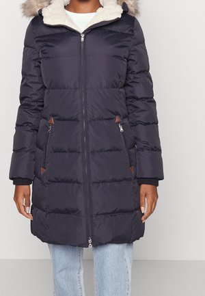 Vrouw draagt een lange zwarte donzen jas met een capuchon met bontkraag, een ritssluiting aan de voorkant en ritssluitingen op de zijzakken, gecombineerd met lichtblauwe spijkerbroek.