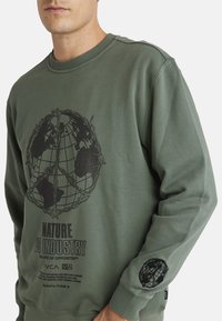 Olive green sweatshirt with a round globe graphic and text in black. Features ribbed neck and cuffs, with logo detail on the sleeve.