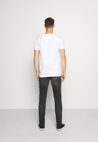 Vit bomulls t-shirt med korta ärmar, parat med mörkgrå slim-fit jeans och svarta sneakers, sedd bakifrån mot en grå bakgrund.