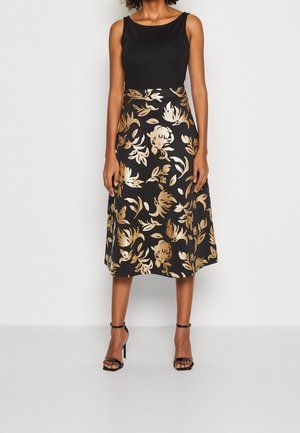 Kobieta ubrana w czarny bezrękawnik i czarną spódnicę midi z złotym kwiatowym wzorem oraz czarne sandały na wysokim obcasie, stojąca na białym tle.