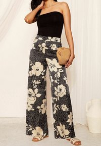 Wide-leg pants in black with large cream floral patterns and white polka dots. Paired with a black strapless top and beige woven clutch.