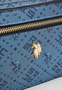 Niebieska torba z tkaniny o wzorze, z złotym zamkiem błyskawicznym i metalowymi elementami, z logo polo gracza w złocie na przodzie, z teksturowanym wzorem.