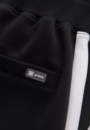 I pantaloni della tuta neri presentano una texture liscia, una striscia di accento bianca e un piccolo etichetta trasparente con la scritta "ombre" sulla tasca laterale.