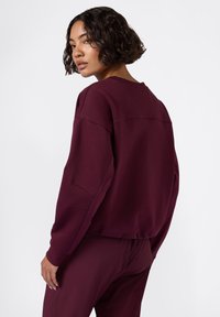 Sudadera burdeos con un corte relajado, puños acanalados y escote de barco. Tejido suave y detalles de costura en la parte posterior. Combinada con pantalones a juego.