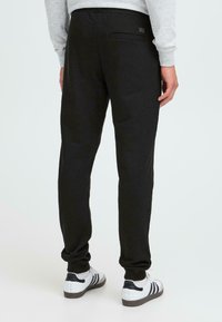 Schwarze Jogginghosen mit schmaler Passform, elastischem Bund und einer einzelnen Gesäßtasche. Getragen mit weißen Sneakers mit schwarzen Streifen.
