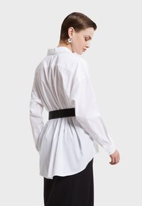 Hvid bomulds skjorte med langeærmer, afslappet pasform, samlet rygdetalje og et sort bælte. Glat tekstur og rundet kant.