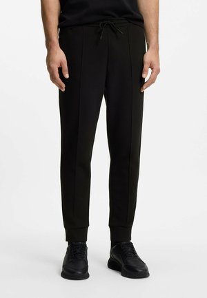 Uomo che indossa pantaloni jogger neri slim fit con cuciture frontali, polsini elasticizzati, vita con coulisse e sneakers nere su sfondo bianco.