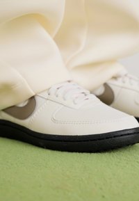 White sneakers with black soles and gray swoosh logos worn with light beige pants, standing on a textured green surface.