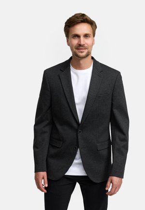Man met lichtbruin haar draagt een zwarte blazer over een wit overhemd en een zwarte broek, staand tegen een effen lichte achtergrond.