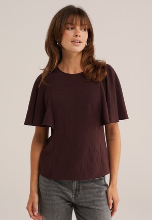 Femme portant un chemisier violet foncé à manches courtes larges et un jean gris, regardant légèrement vers sa gauche sur un fond uni.