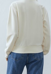 Sudadera de color crema con una textura suave, corte relajado y ribete acanalado, combinada con jeans de mezclilla azul que presentan detalles de costura sutiles.