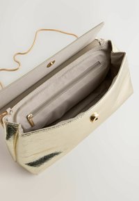 Pochette métallique dorée avec rabat ouvert, montrant un intérieur en tissu beige et une poche intérieure zippée, ainsi qu'une fine chaîne dorée.