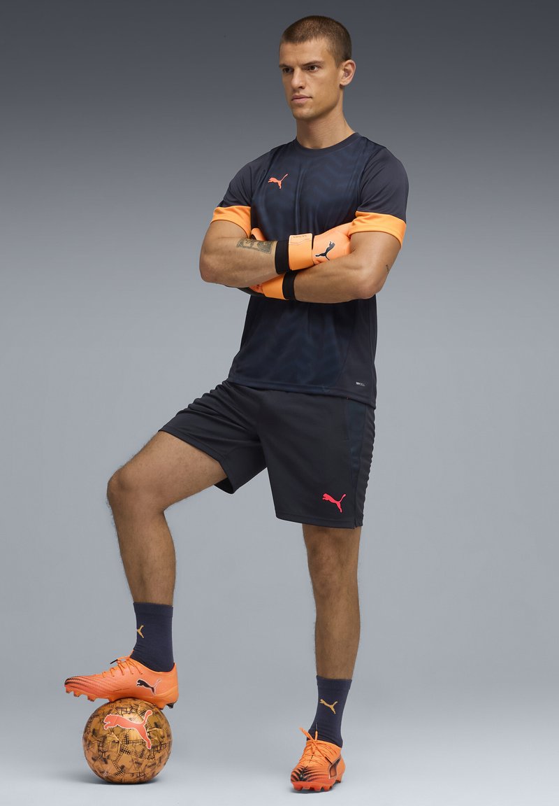 La indumentaria atlética de Puma presenta una parte superior de un azul marino oscuro con mangas naranjas, combinada con pantalones cortos azul marino. El jugador lleva unos zapatos de fútbol de un naranja brillante y sostiene un balón de fútbol.