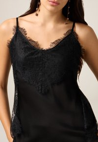 Schwarzes Unterhemd mit zarter Spitzenverzierung entlang des Ausschnitts und der Schultern, aus glattem, halbtransparentem Stoff mit verstellbaren Trägern.