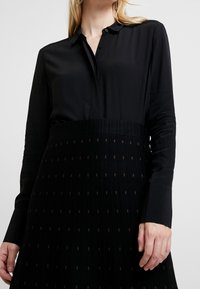 Chemise noire à manches longues avec col, présentant une texture lisse, associée à une jupe plissée noire affichant un motif à pois subtil.