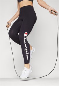 Mustat korkeavyötäröiset legginssit, joissa on valkoinen "Champion"-logo ja punainen yksityiskohta, sileä kangas, yhdistetty valkoisiin urheilukenkihin ja hyppynaruun.