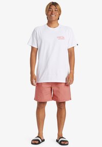 Biela bavlnená tričko s logom "Quiksilver"; koralové šortky z ľahkého materiálu; čierne sandále s bielymi pruhmi na popruhoch.