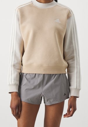Sweat-shirt Adidas beige et blanc cassé avec trois bandes sur les manches, associé à un short de sport Adidas gris clair.