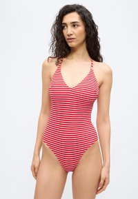 Γυναίκα με μακριά σγουρά μαλλιά που φοράει ολόσωμο μαγιό με κόκκινες και λευκές ρίγες, κοιτάζοντας προς το πλάι, στέκεται μπροστά σε απλό φόντο.