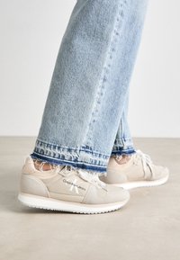 Beżowe trampki Calvin Klein Jeans mają teksturowaną cholewkę, białą podeszwę i sznurowadła, zestawione z jasnoniebieskimi jeansami zniszczonymi na dole.