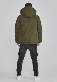 Olivenfarvet puffjakke med stor hætte, justerbare stropper og synlige sømme. Båret med slim-fit sorte cargo bukser og hvide sneakers.