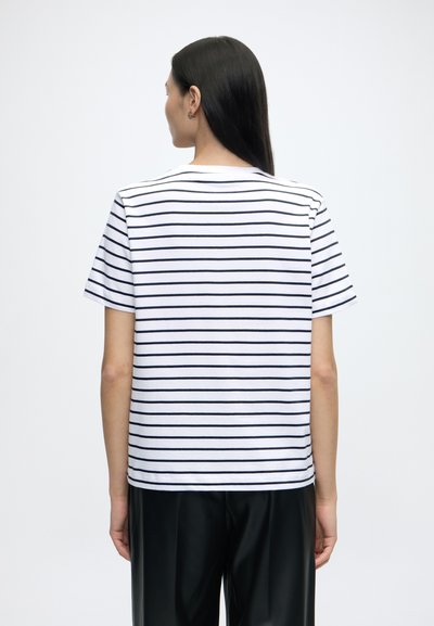 Femme aux longs cheveux foncés portant un t-shirt blanc à manches courtes avec de fines rayures horizontales noires, de dos, sur un fond clair uni.