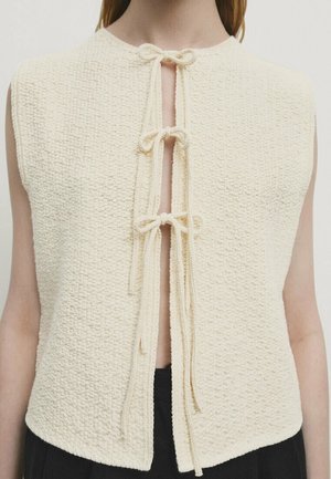 Bezrękawnik w kremowym kolorze o fakturowanej strukturze z trzema zawiązanymi kokardami z przodu, noszony na jasnej karnacji i zestawiony z czarnymi spodniami.