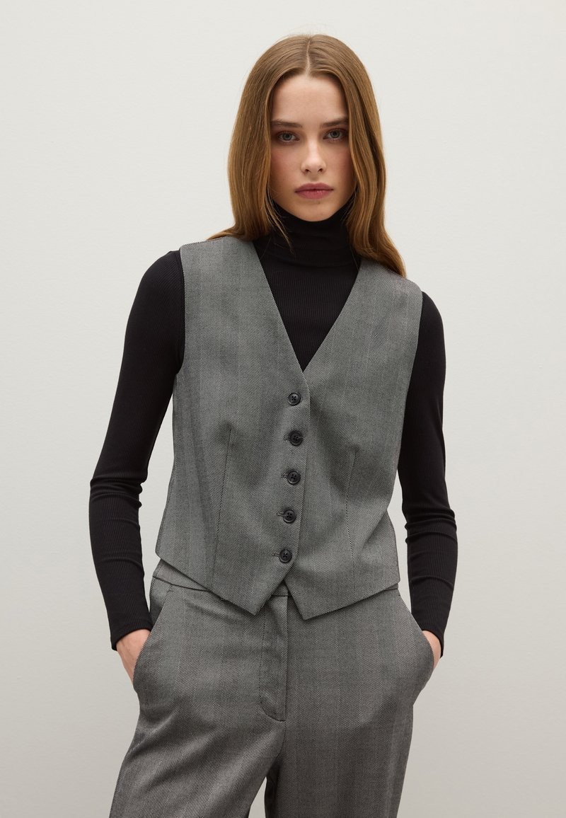 Gilet grigio a micro motivo houndstooth con bottoni, abbinato a un dolcevita nero. Design slim-fit, forma strutturata, tessuto testurizzato e tasche laterali.