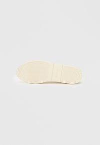 Semelle en caoutchouc beige clair avec des nervures texturées pour une meilleure adhérence, comportant une zone en creux avec un logo en relief et un contour lisse.