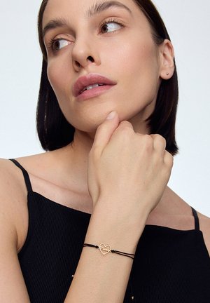 Złota bransoletka w kształcie serca na czarnejCord, z napisem "Mum". Modelka nosi czarny top na ramiączkach, na neutralnym tle i o jasnej karnacji.