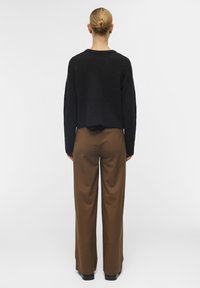 Suéter negro cortado con tejido texturizado, combinado con pantalones marrones de corte holgado. Zapatos oscuros completan el conjunto.
