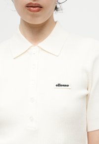 Femme portant un polo blanc à manches courtes avec des boutons et un patch logo "ellesse" sur la poitrine.