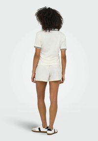 Weißes Baumwollpolo mit schwarz gestreiftem Kragen, kombiniert mit passenden Shorts. Das Modell trägt weiße Sneakers mit schwarzen Akzenten und Gummisohlen.