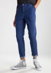 Modré džíny s ohrnutými nohavicemi, s knoflíkovým zapínáním a předními kapsami, kombinované se šedými vysokými teniskami.