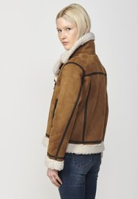 Giacca in suede marrone con colletto e polsini in shearling crema, caratterizzata da dettagli in piping nero e un design aderente.